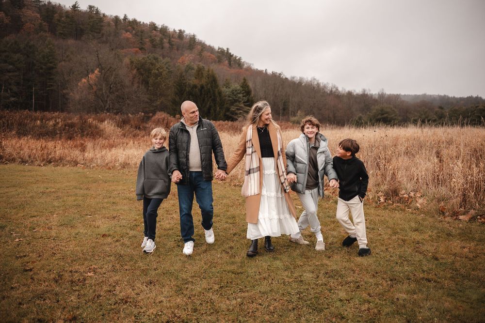 A family stands together in a grassy field with autumn foliage and mountains in the background.