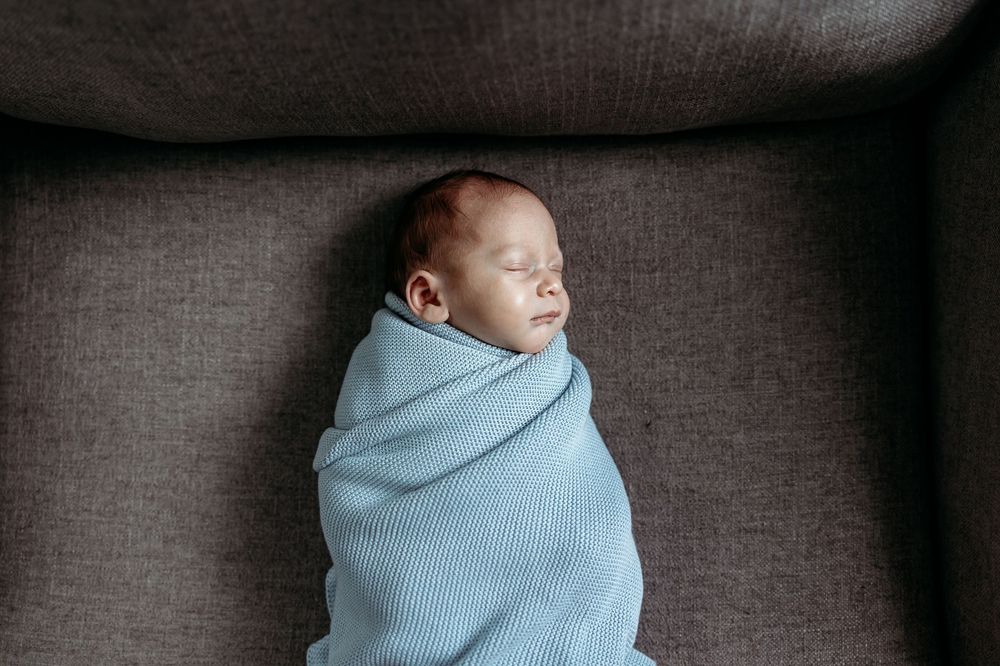 A sleeping newborn swaddled in a soft blue blanket rests peacefully on a gray couch.