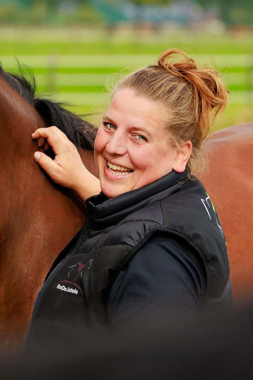A person in a black jacket smiles while petting a brown horse on an outdoor field.