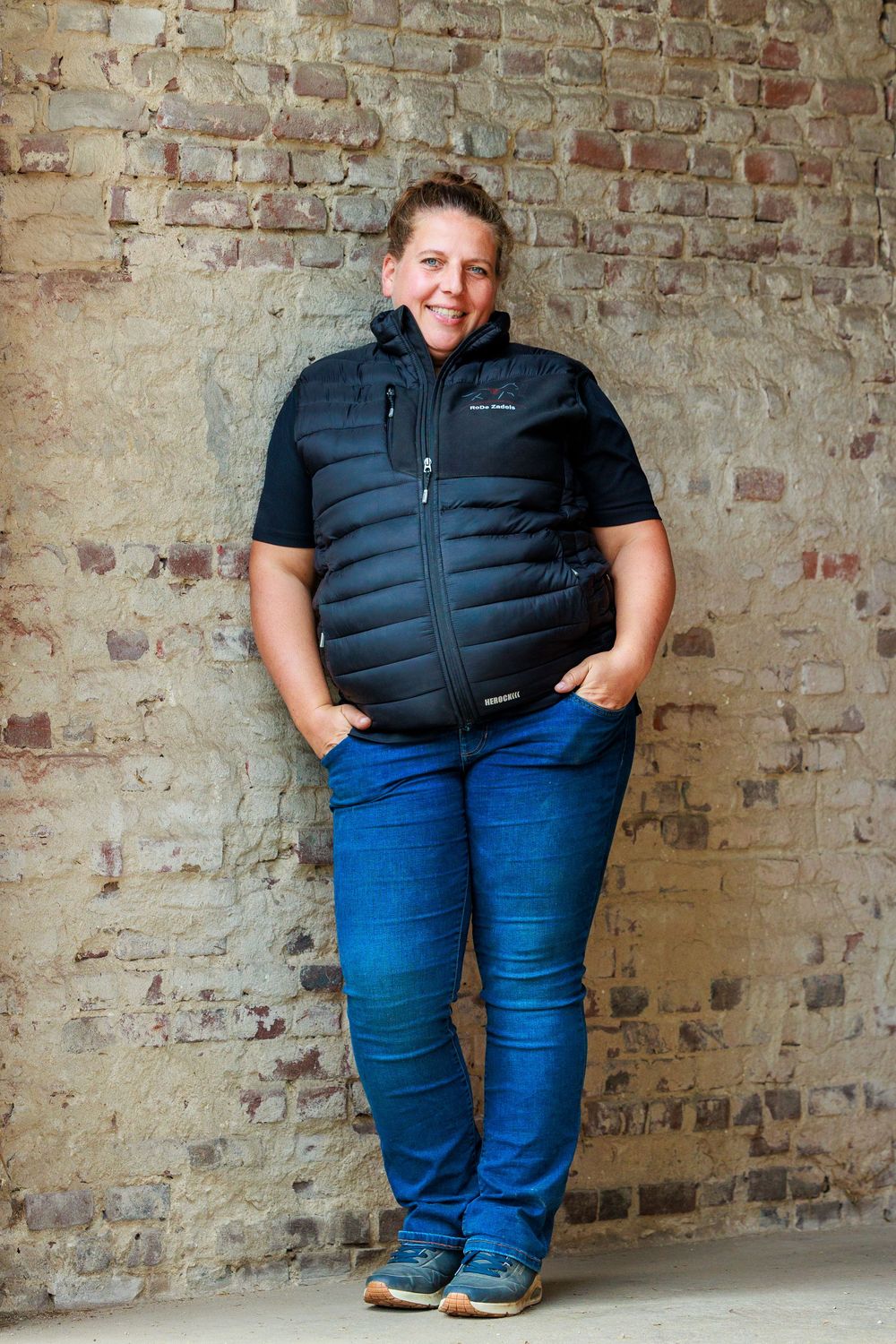 Person wearing a black puffer vest and blue jeans poses against a rustic brick wall in a photo series.