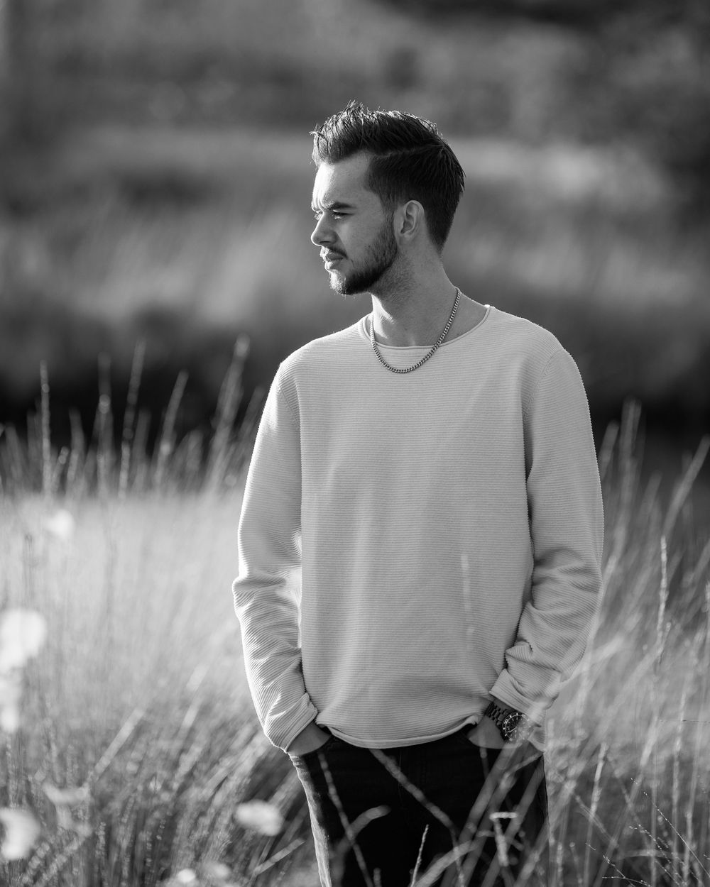 A person wearing a white sweater sits thoughtfully in a grassy field on a sunny day, captured in black and white.