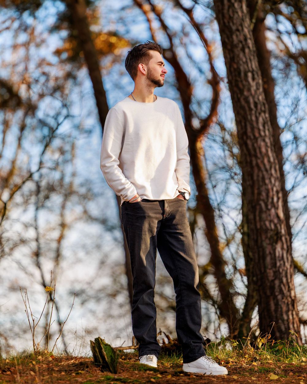 Person in a white sweater and dark pants poses thoughtfully against autumn trees in a scenic outdoor setting.