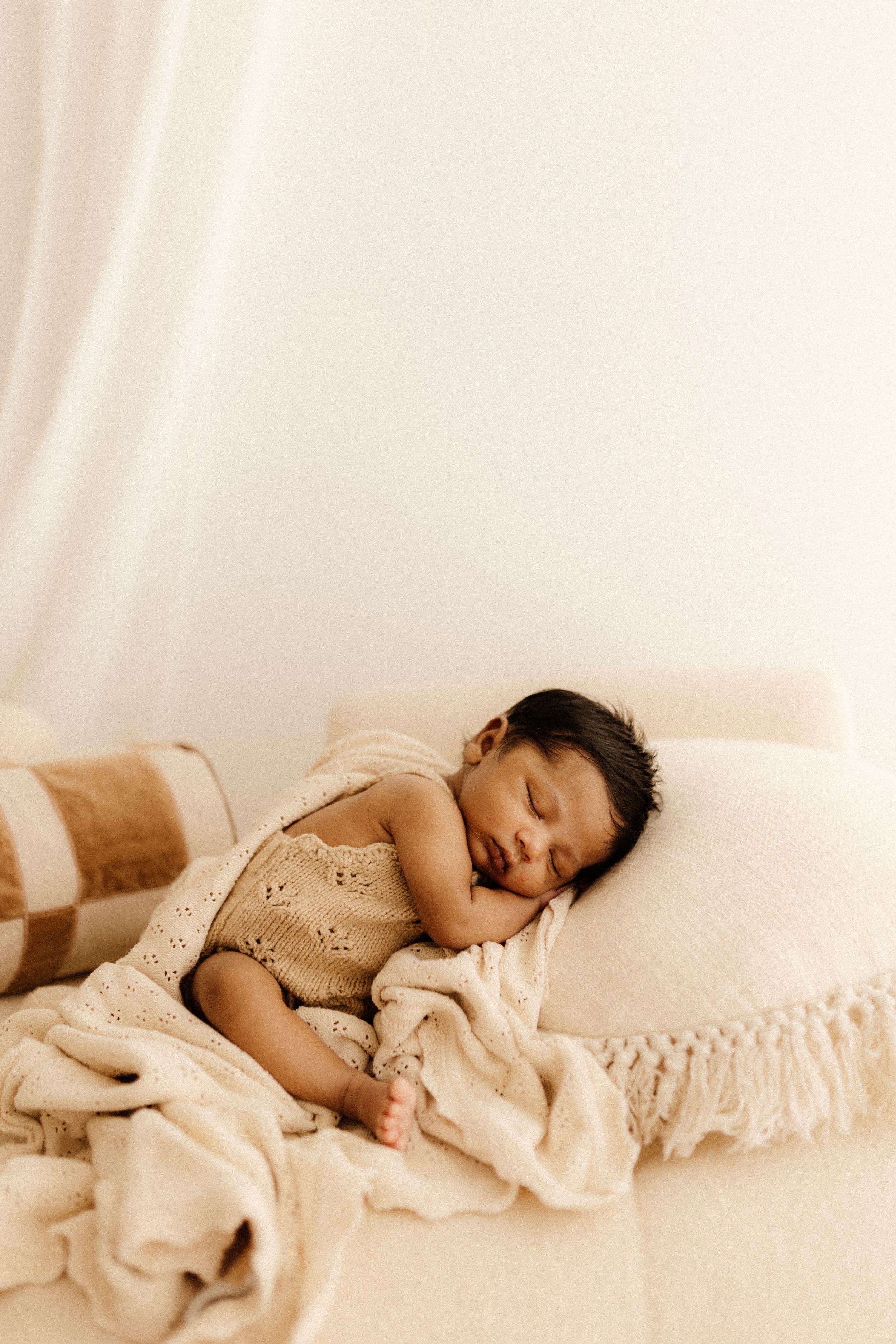 Sleeping baby in textured outfit rests on cream couch with fringe throw blanket.