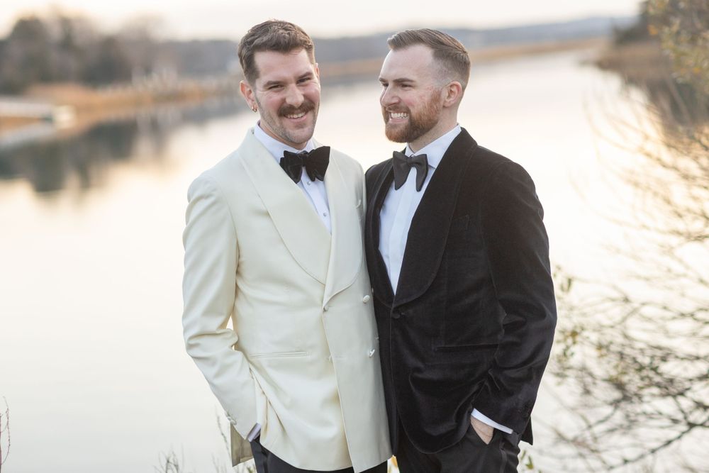 Two grooms in tuxedos smiling together by a reflective lake at sunset.
