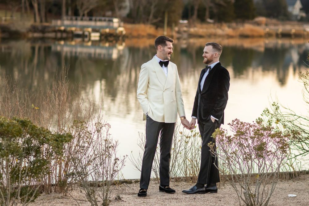 Two grooms in formal attire holding hands by a serene lake at sunset.