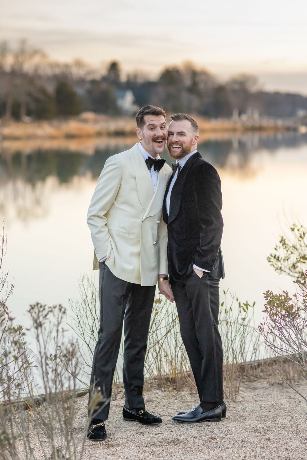 Two grooms laughing together in tuxedos by a peaceful lake at sunset.