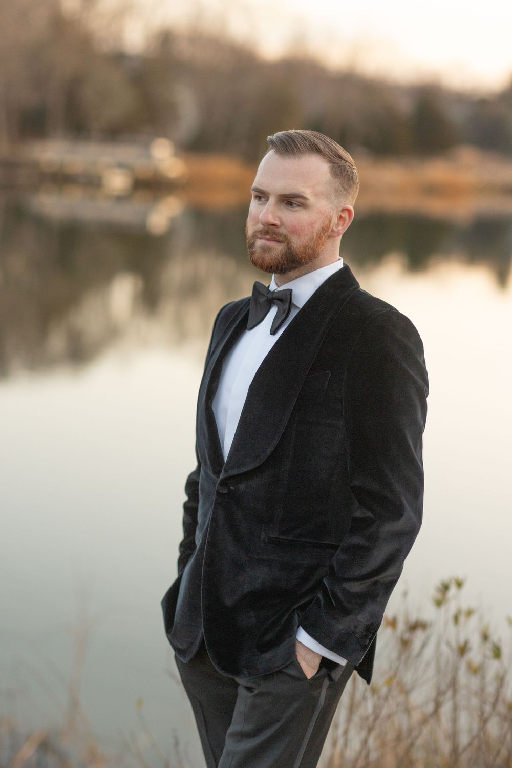 Groom in black velvet tuxedo posing confidently by a calm reflective lake.