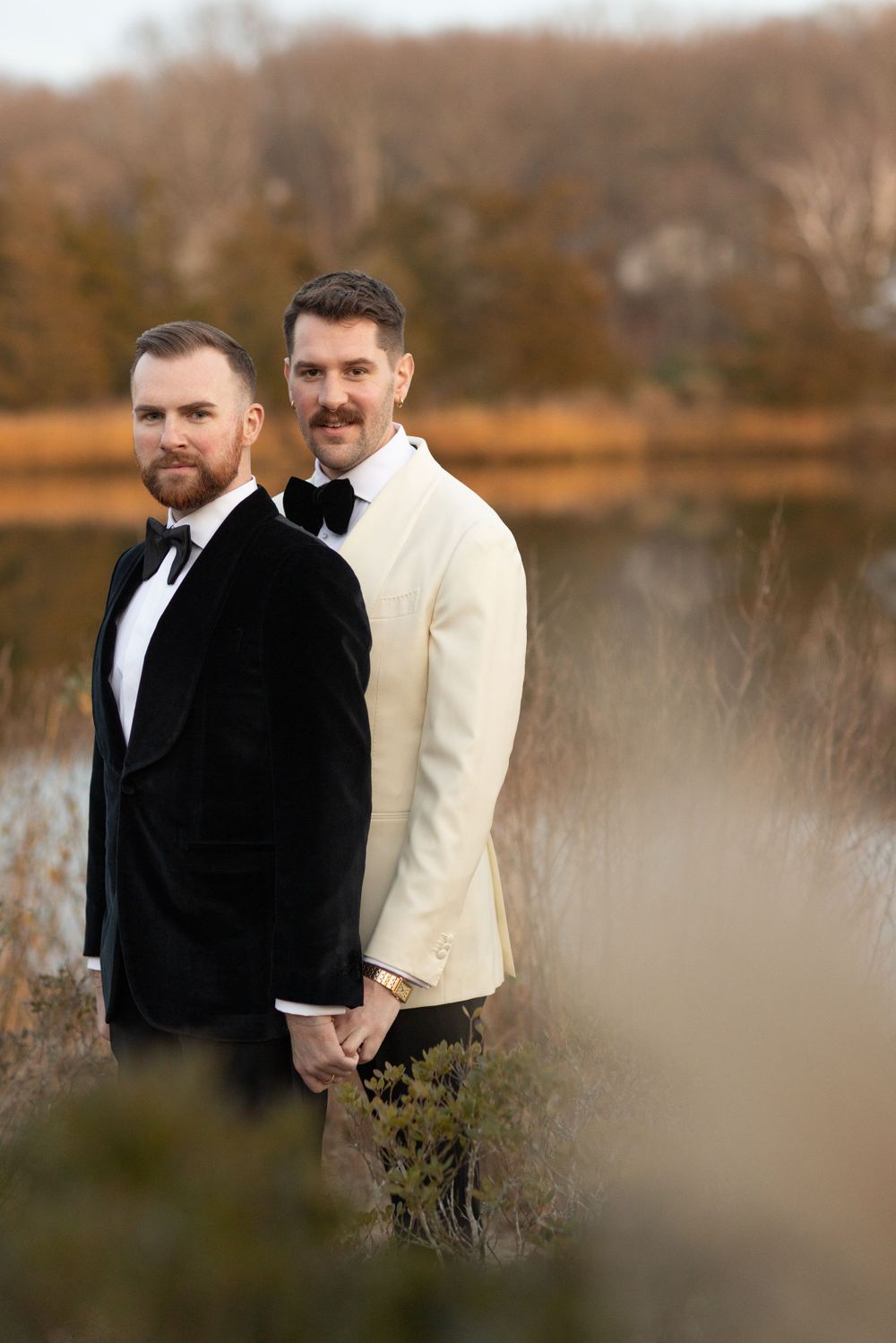 Two grooms in black velvet and ivory tuxedos hold hands outdoors near a lake during their wedding.