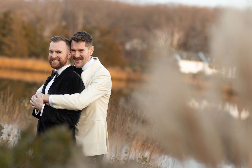 Two grooms embrace and smile outdoors near a lake in black and ivory wedding tuxedos.