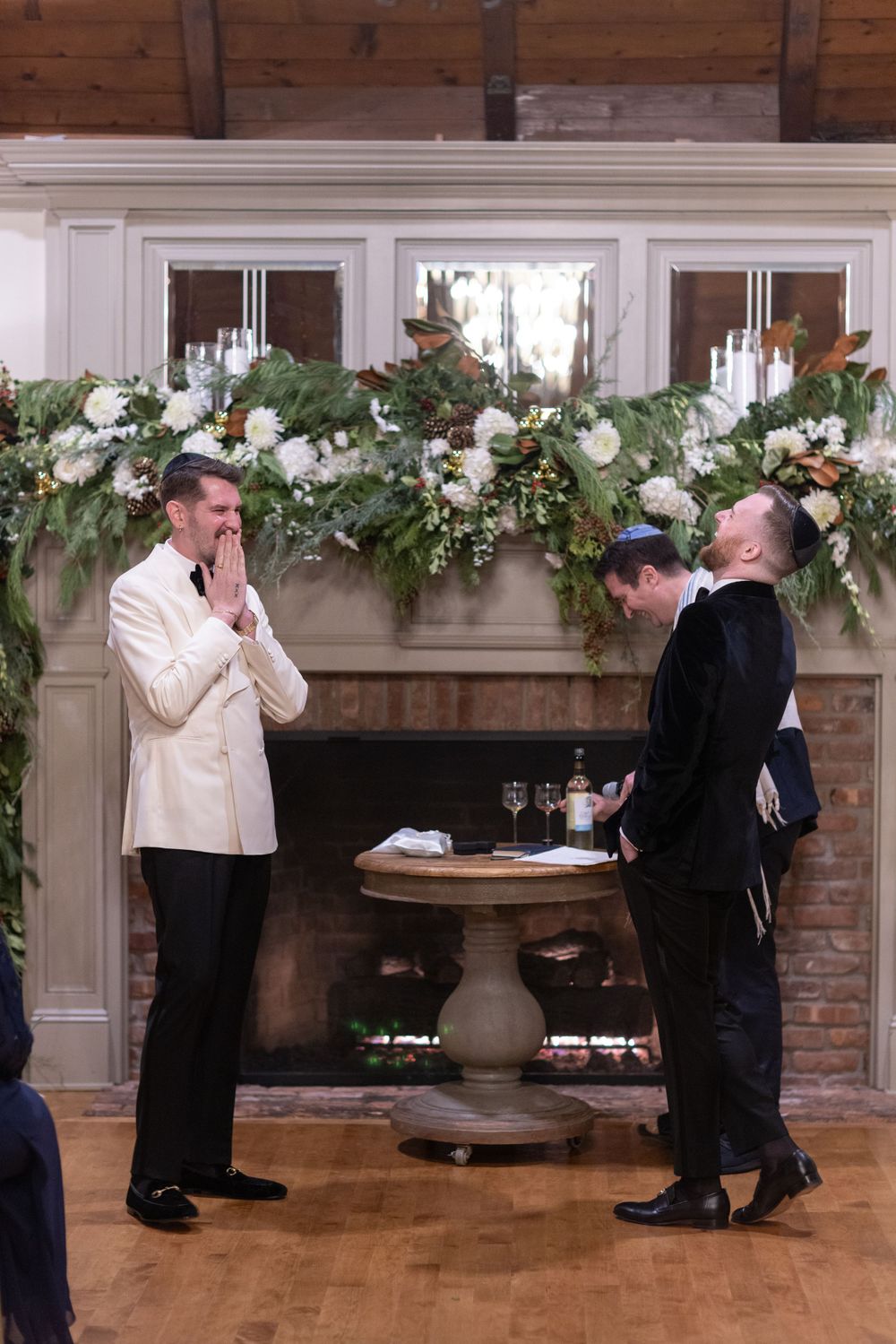 Two grooms share a joyful moment with officiant at Jewish wedding ceremony by decorated fireplace.