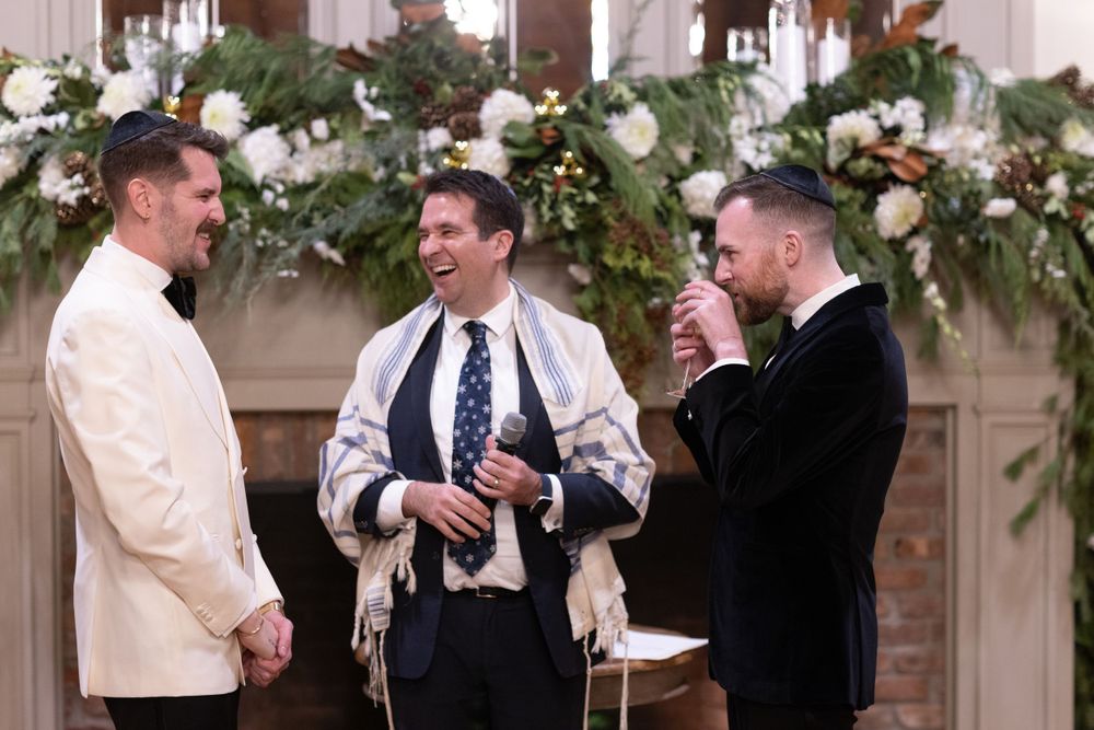 Two grooms laugh with Jewish officiant wearing tallit at elegant indoor wedding ceremony.