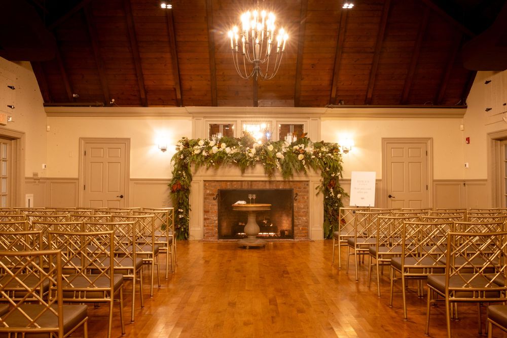 Elegant wedding ceremony venue with gold chiavari chairs and a floral greenery arch over a brick fireplace.