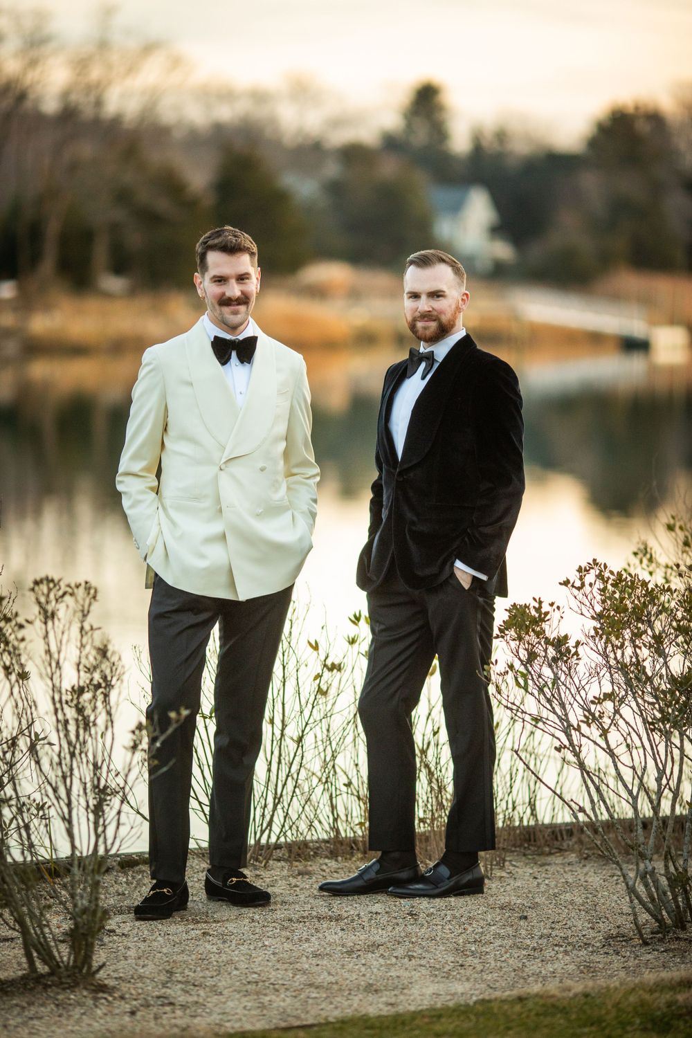 Two grooms in formal attire pose by a scenic lakeside at sunset during their wedding celebration.