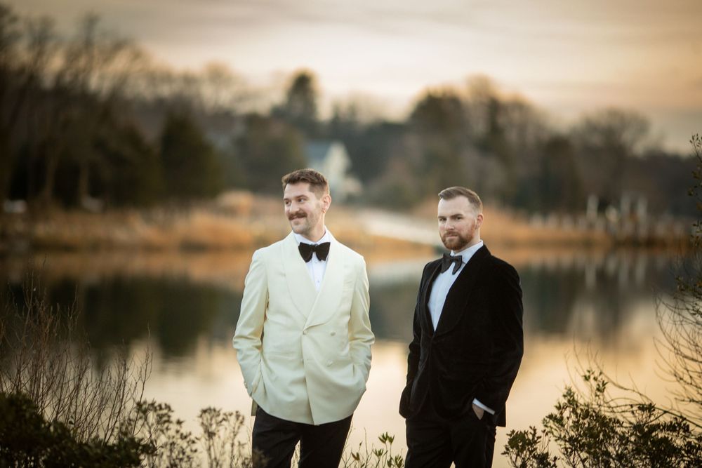 Two grooms in black and ivory tuxedos share a candid moment by a tranquil lake at sunset.