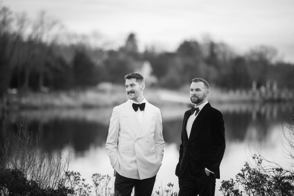 Two grooms in black and white tuxedos pose by a lakeside in a classic black and white wedding portrait.