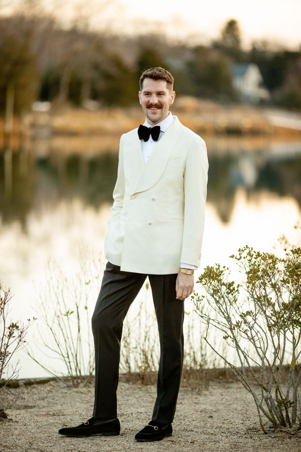 Groom in ivory double-breasted tuxedo jacket with black bow tie poses by a serene lakeside at sunset.