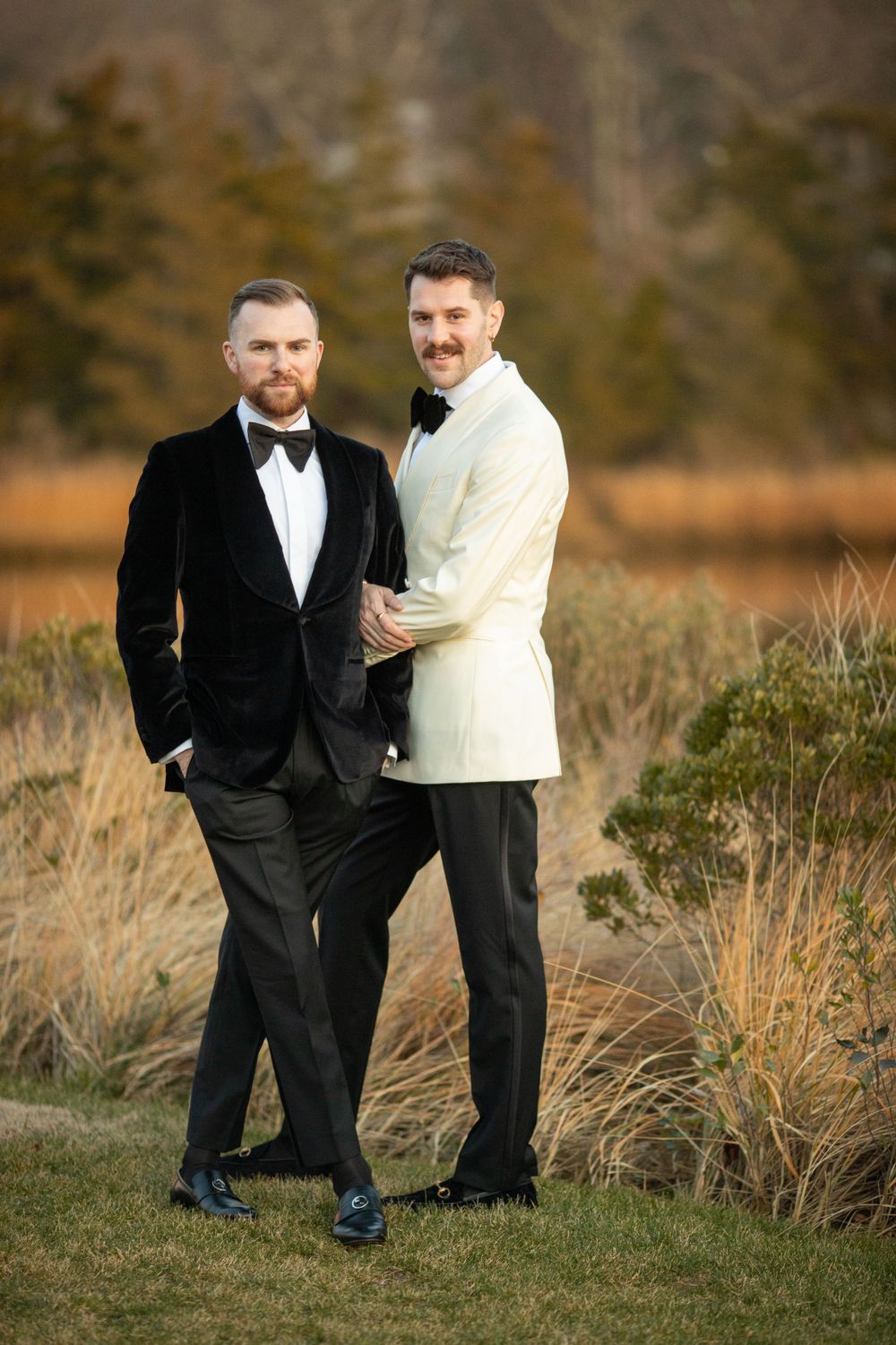 Two grooms, one in black velvet tuxedo and one in ivory jacket, pose together in an outdoor wedding portrait.