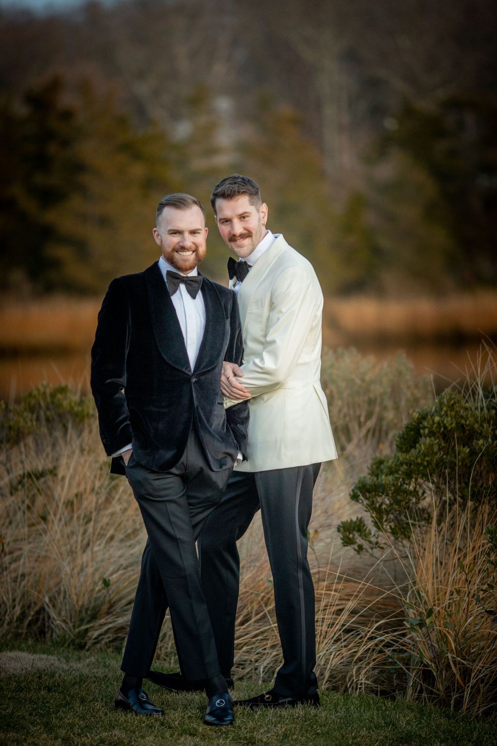Two grooms smiling together outdoors, one in black velvet tuxedo and one in white jacket, with autumn reeds behind.
