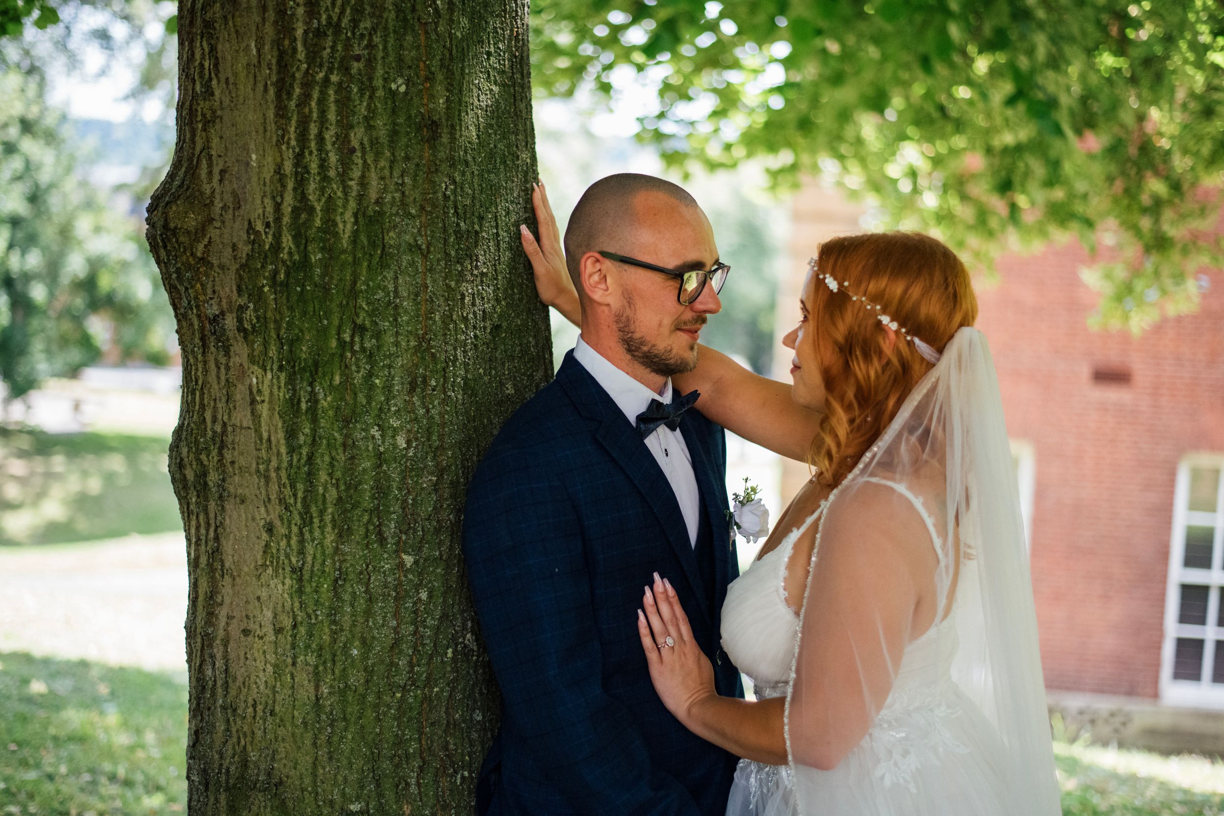 Tender moment between couple under green tree canopy during wedding photoshoot.