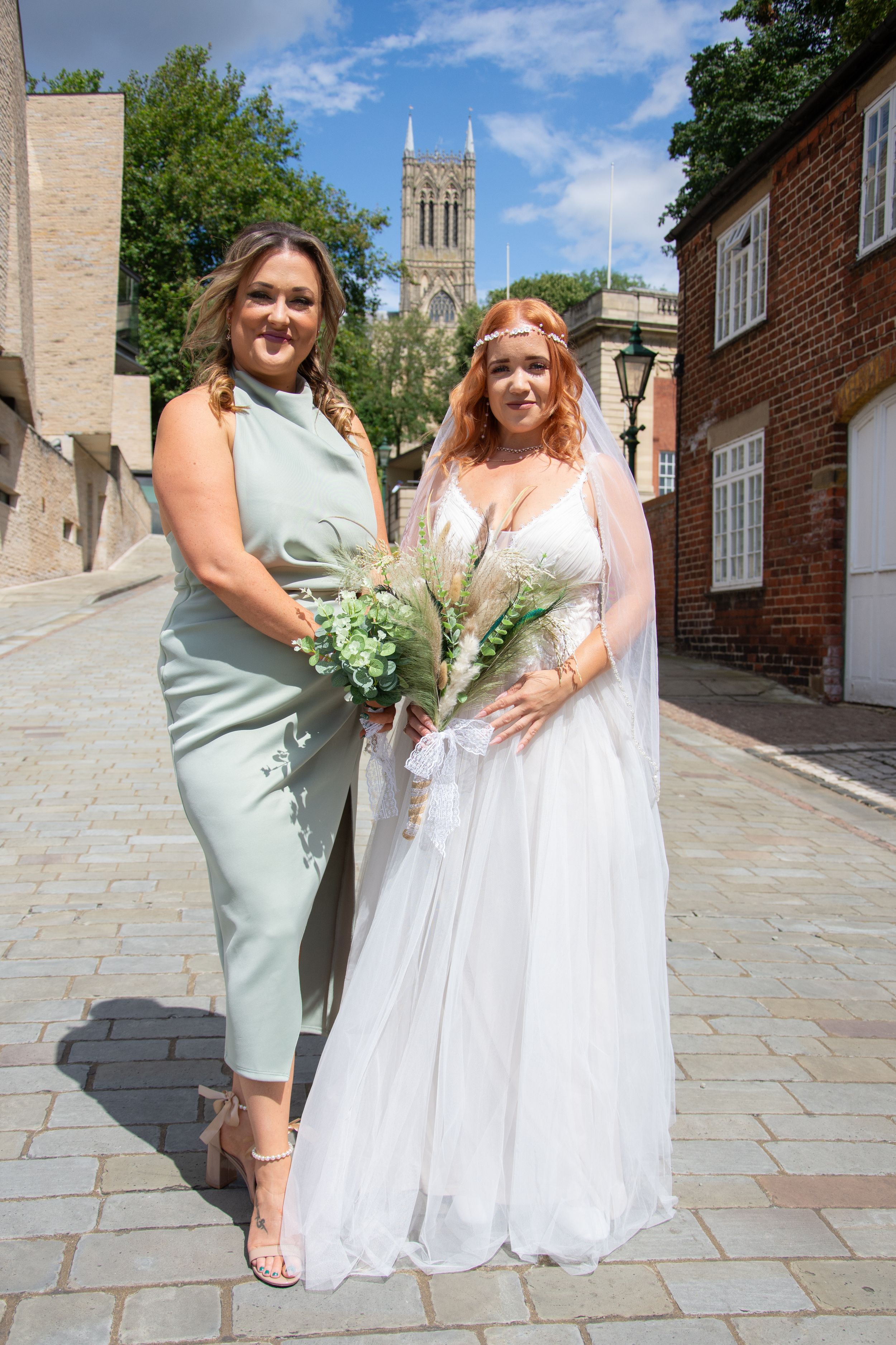 Two people in formal attire standing on a cobblestone path with a cathedral visible in the background.