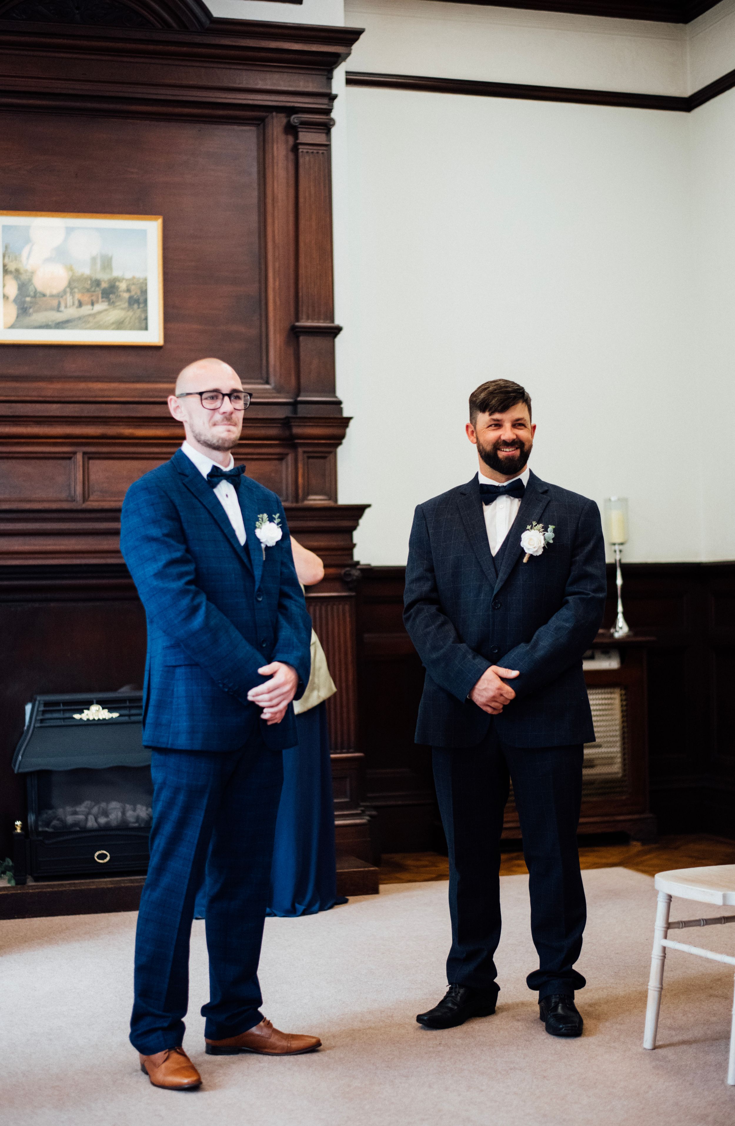 Two people in formal navy suits standing in elegant wood-paneled room.
