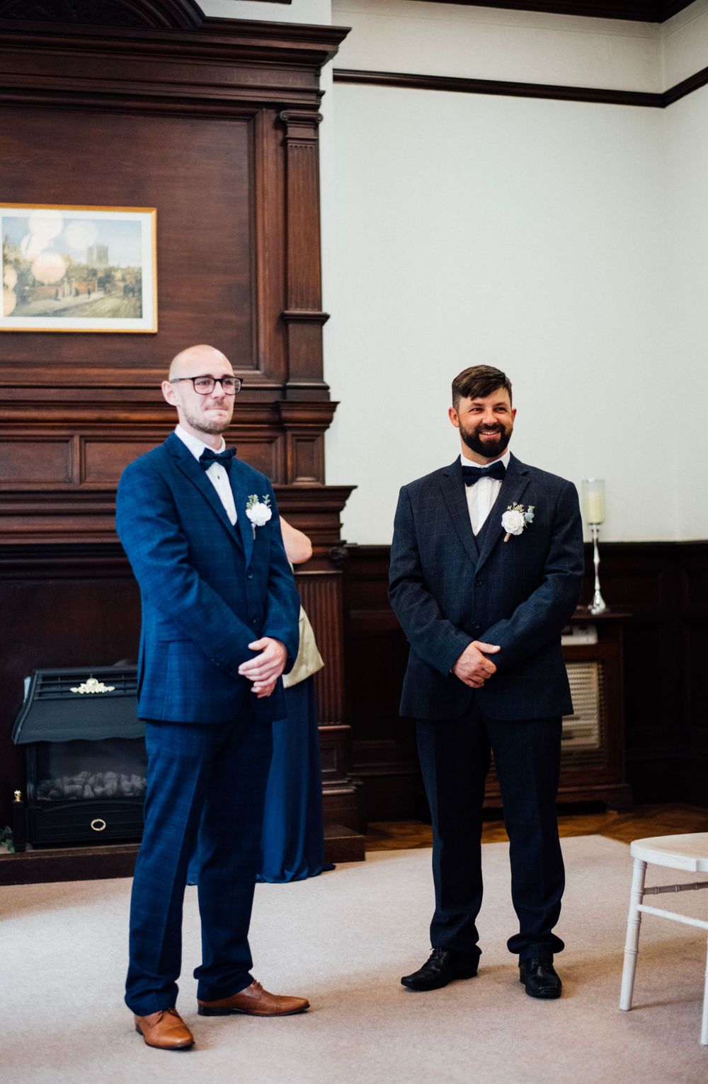 Two people in formal navy suits standing in elegant wood-paneled room.