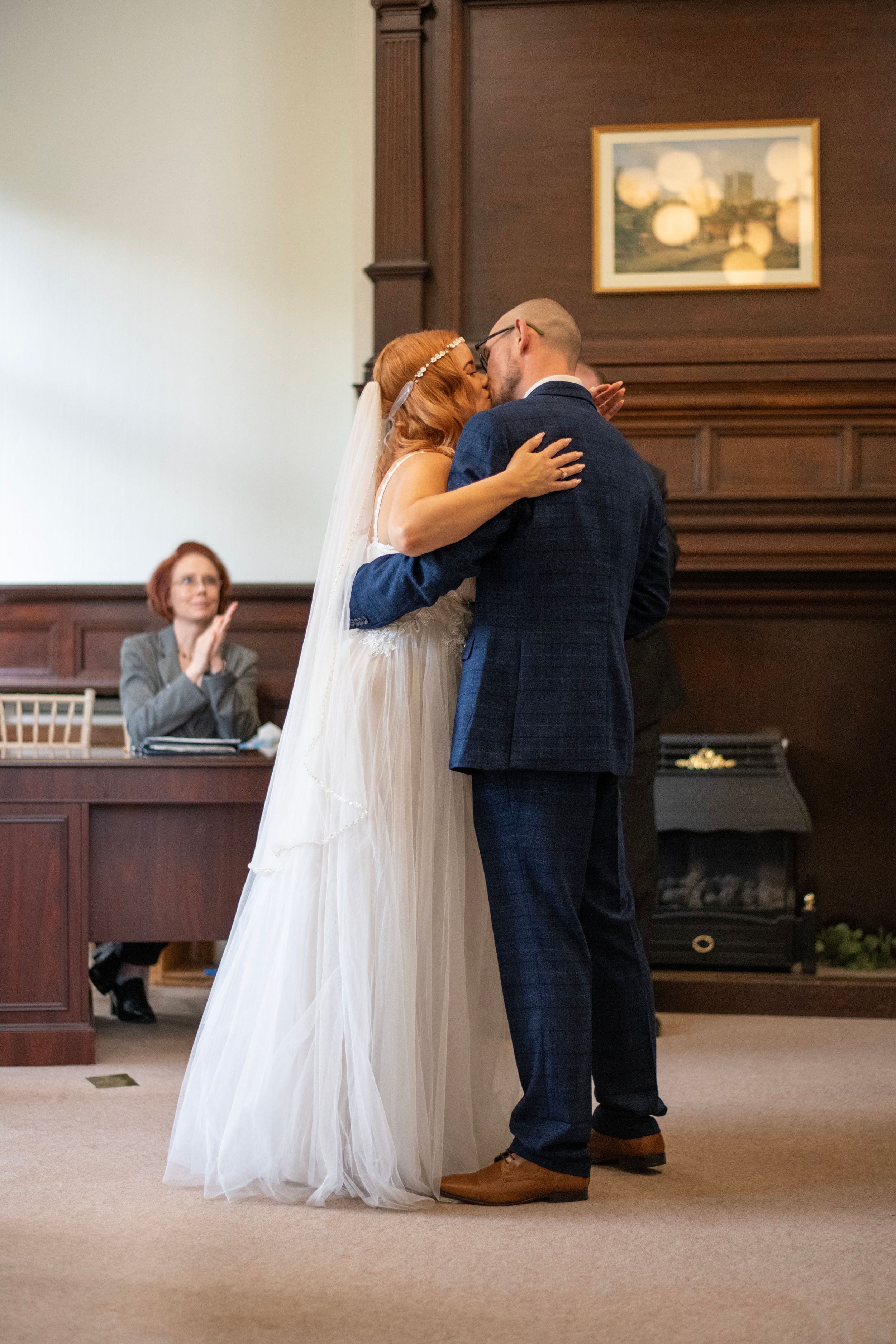 Wedding ceremony first kiss in elegant wood-paneled room with witness in background.