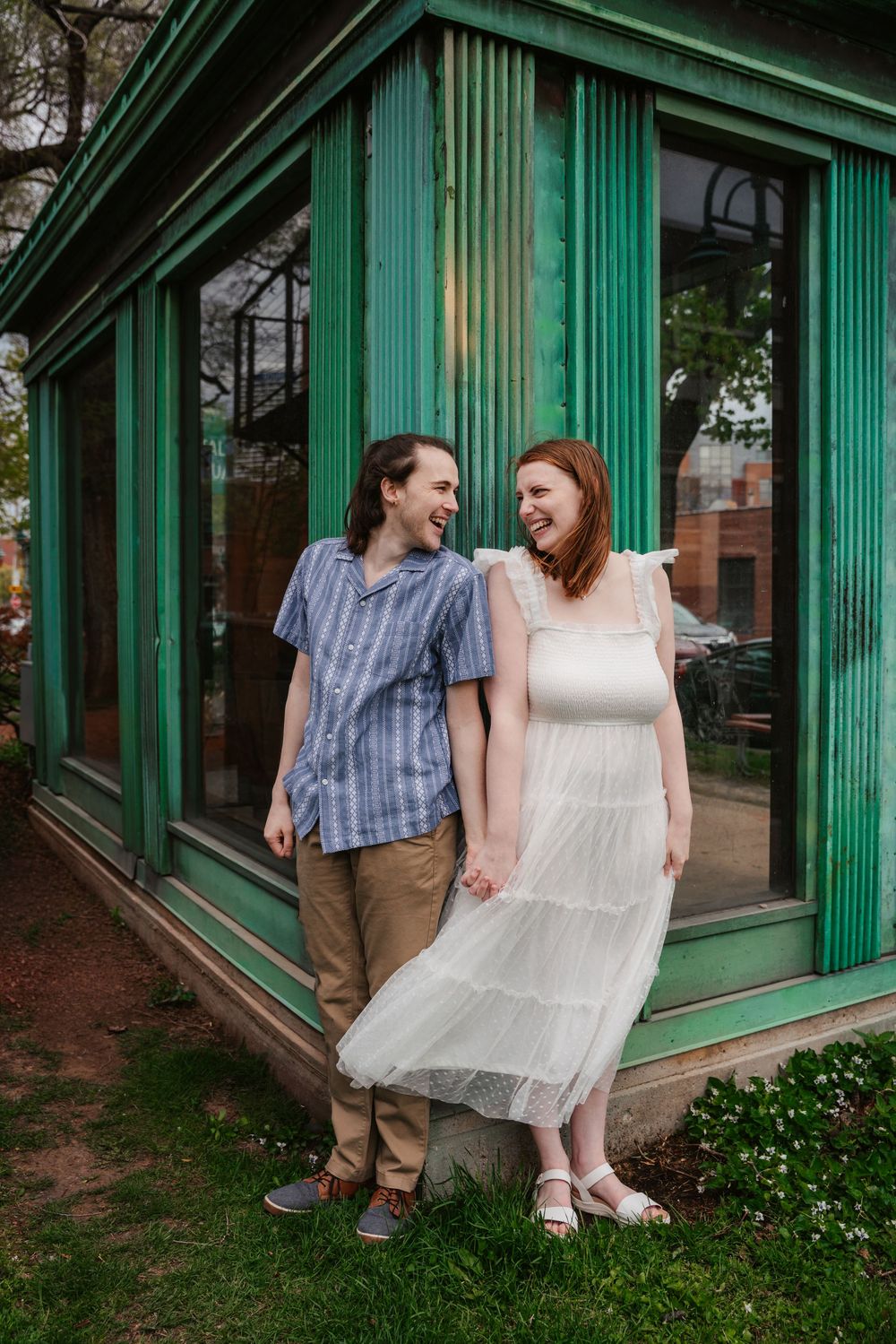 A laughing couple holds hands in front of a green building, she in a white dress, he in a blue shirt and khakis.