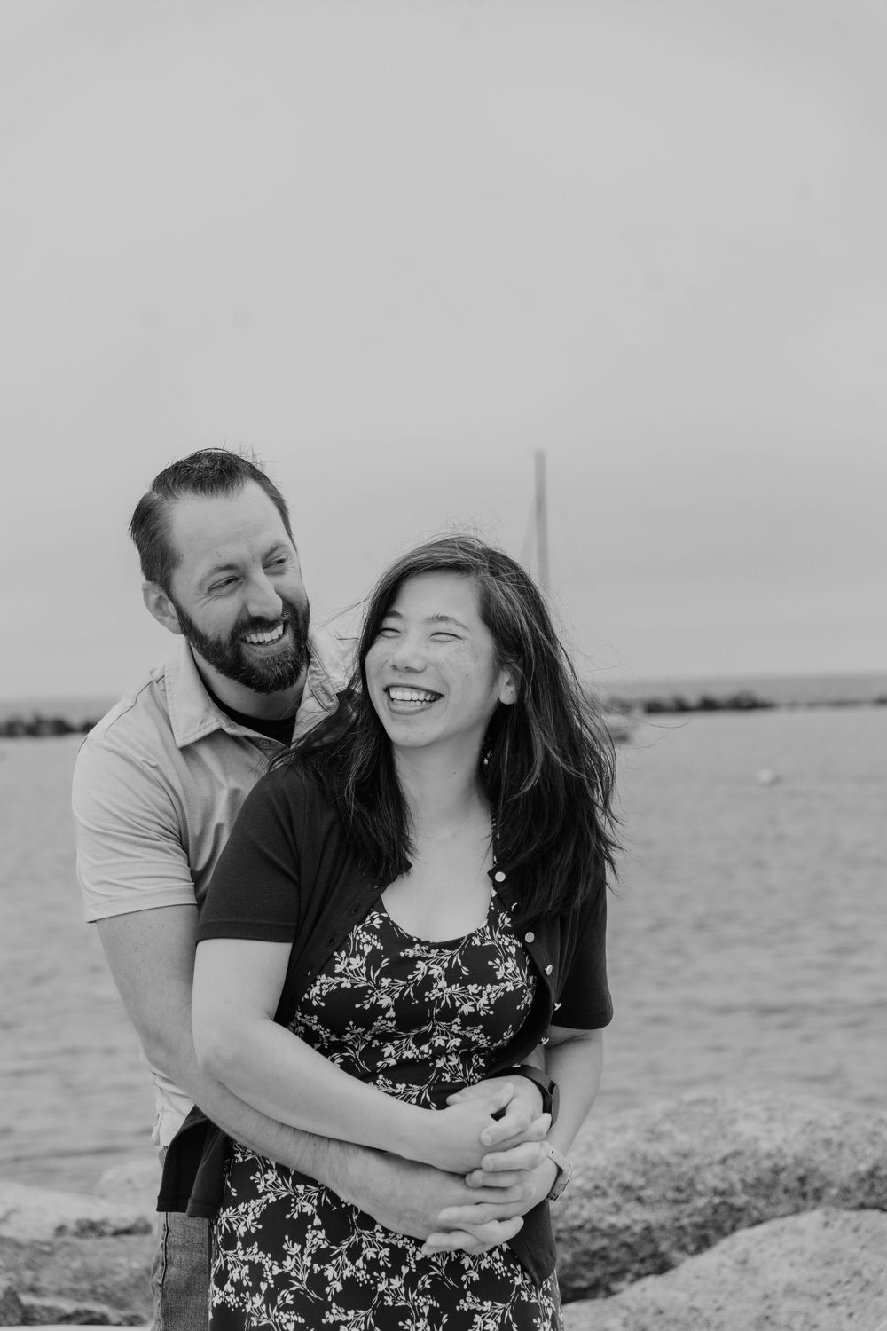 A laughing couple embraces near a rocky shoreline in a black and white outdoor portrait photo.