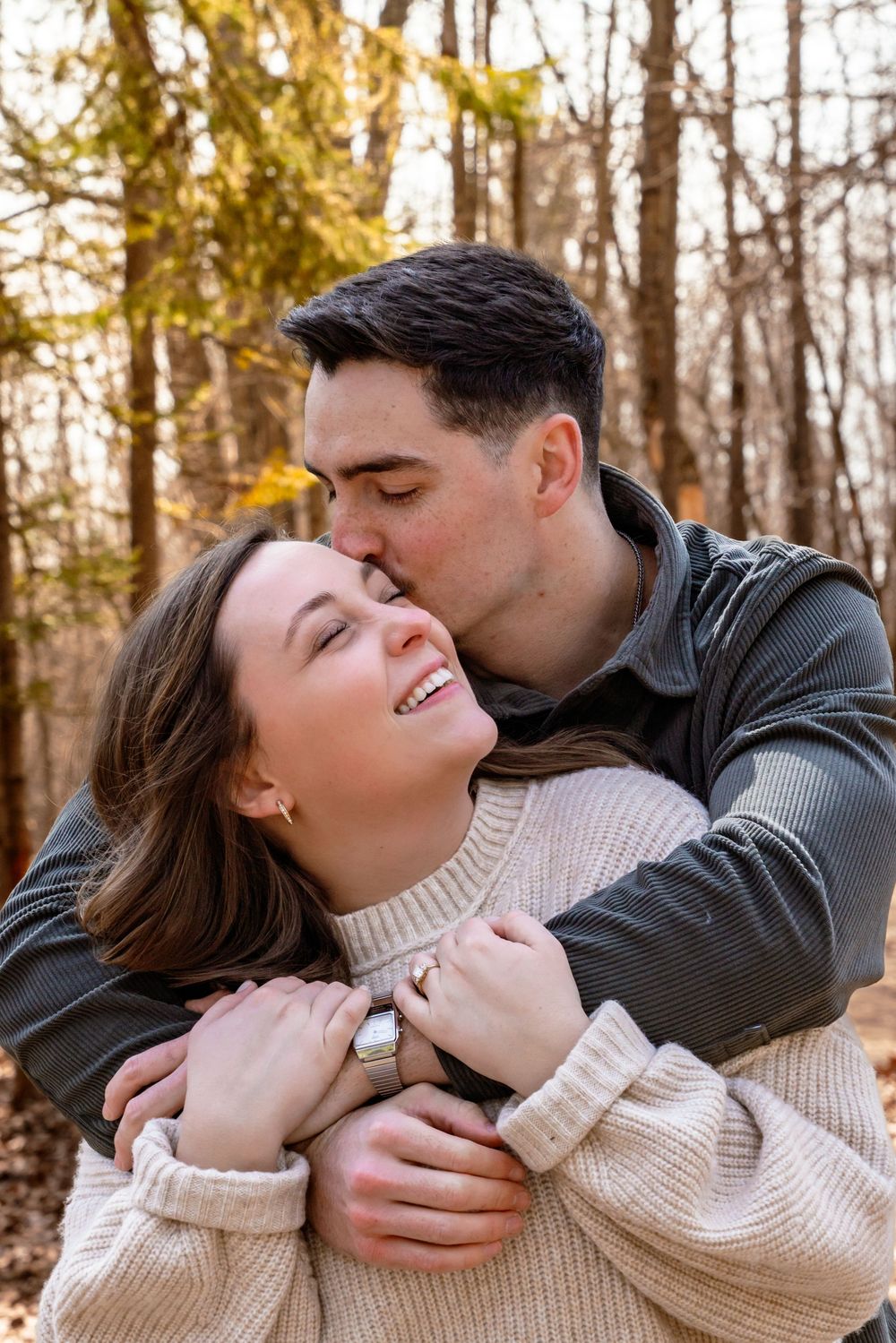 A man kisses a smiling woman on the cheek while embracing her from behind in an autumn forest setting.