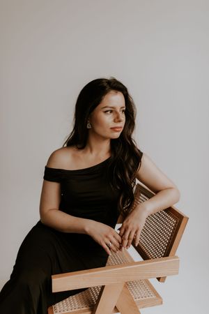 Woman in black dress poses artistically with wooden chair against white wall.