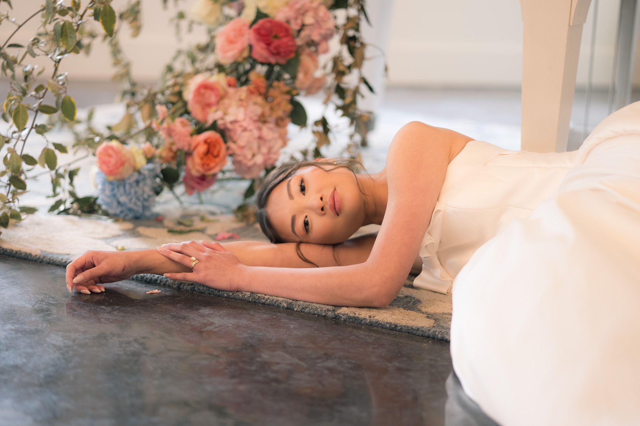 Bride in white gown lying gracefully on marble floor with pink and coral floral arrangement in background.