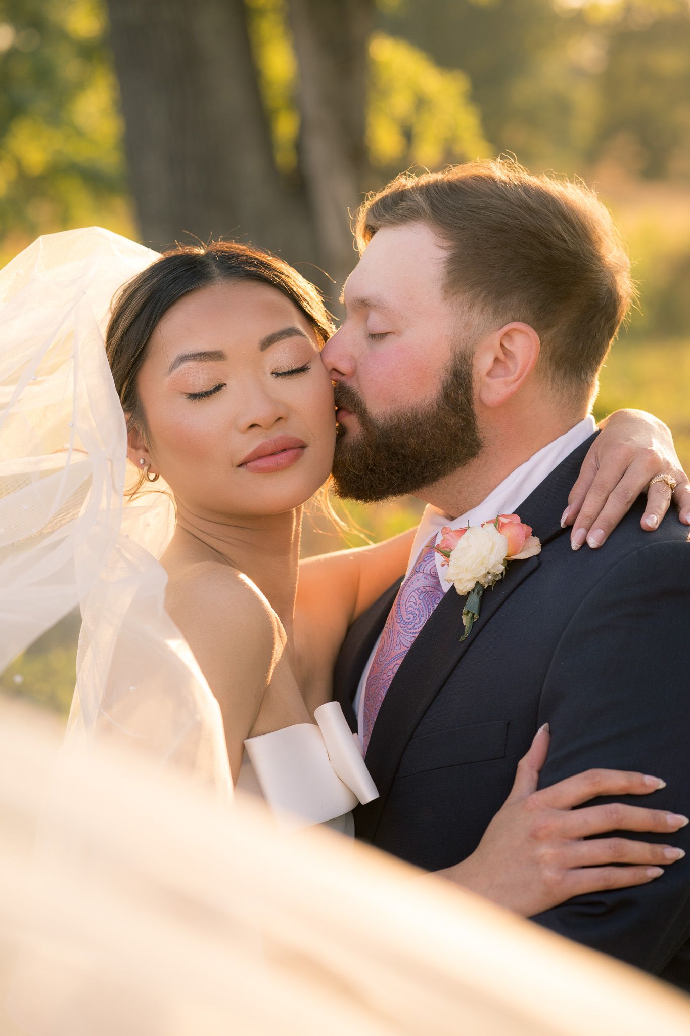 Groom with beard kisses bride wearing veil in golden sunset light outdoors.