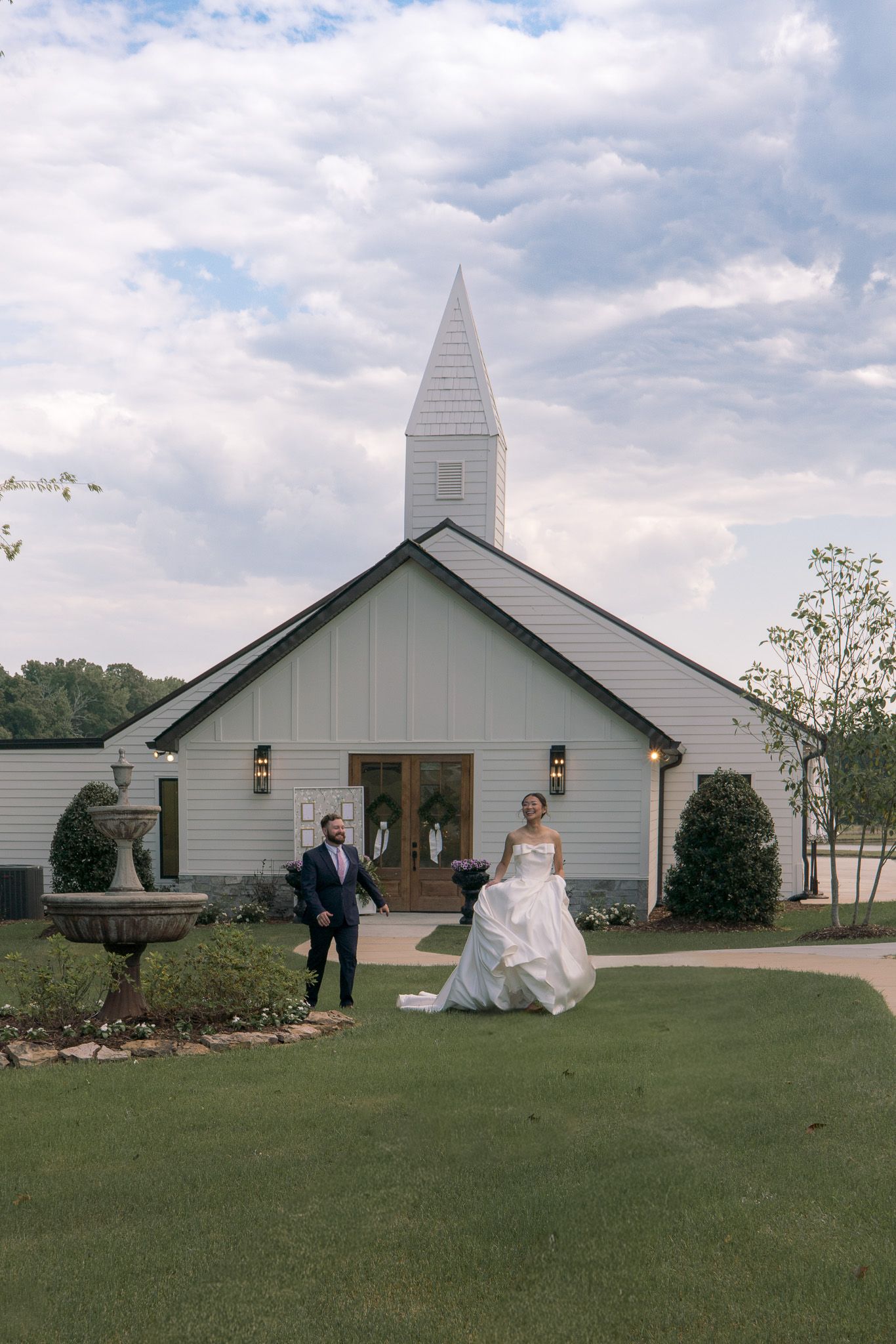 Bride and groom walking outside a white chapel wedding venue with a steeple, lush green lawn, and cloudy sky backdrop.