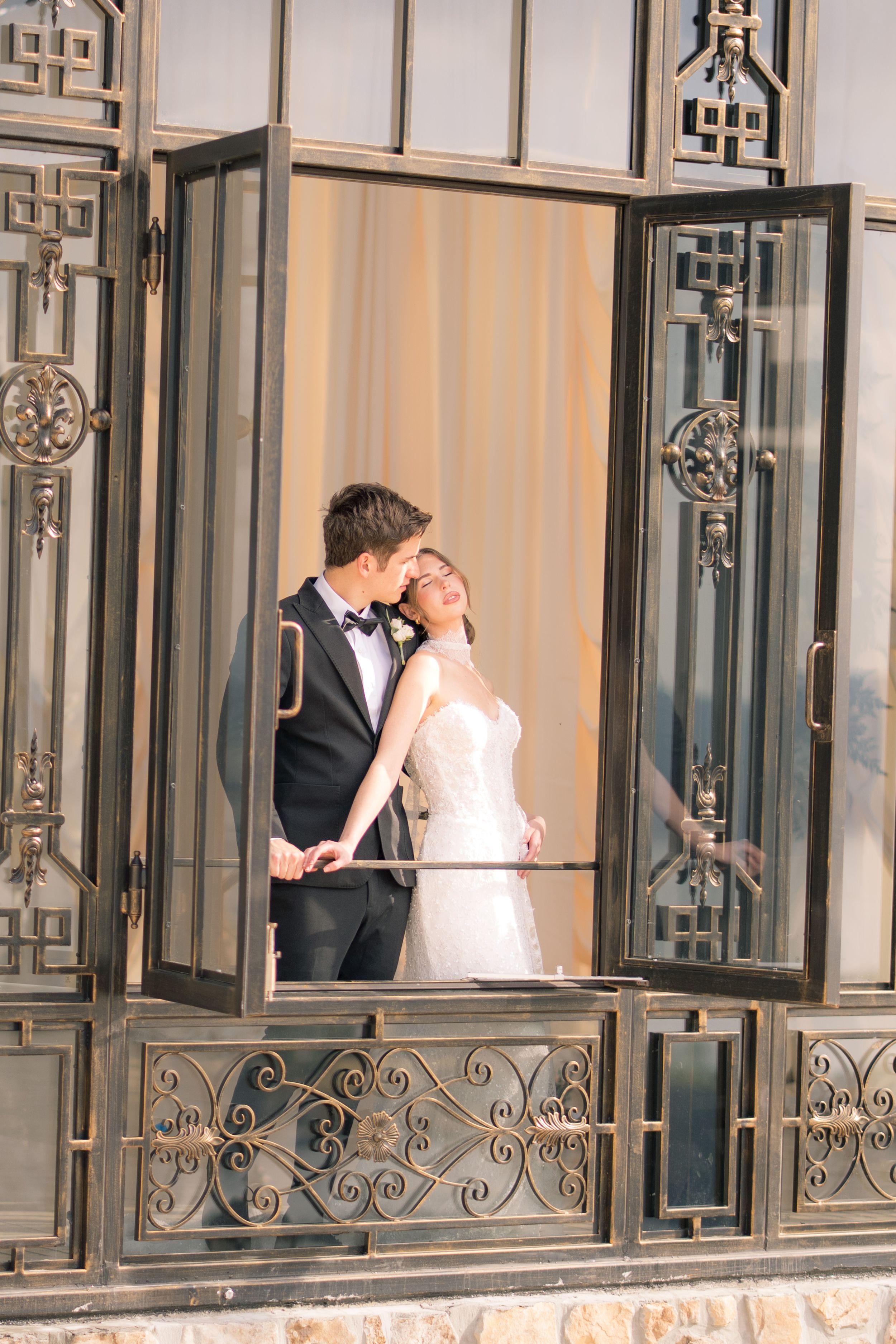 Bride and groom share a romantic moment on an ornate balcony, with the bride in a white gown and groom in a tuxedo.