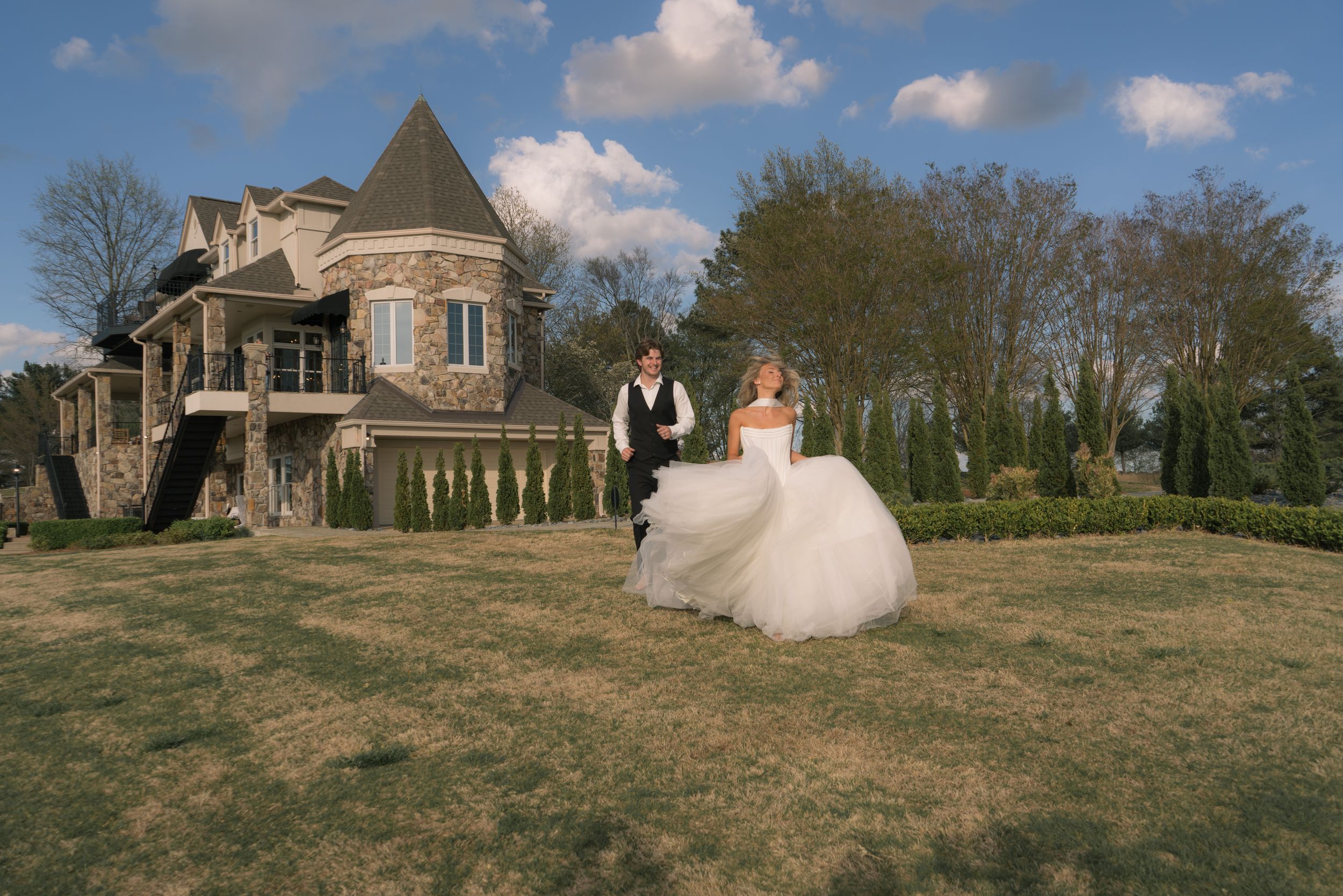 Bride and groom holding hands running across lawn in front of elegant stone castle venue on sunny day.