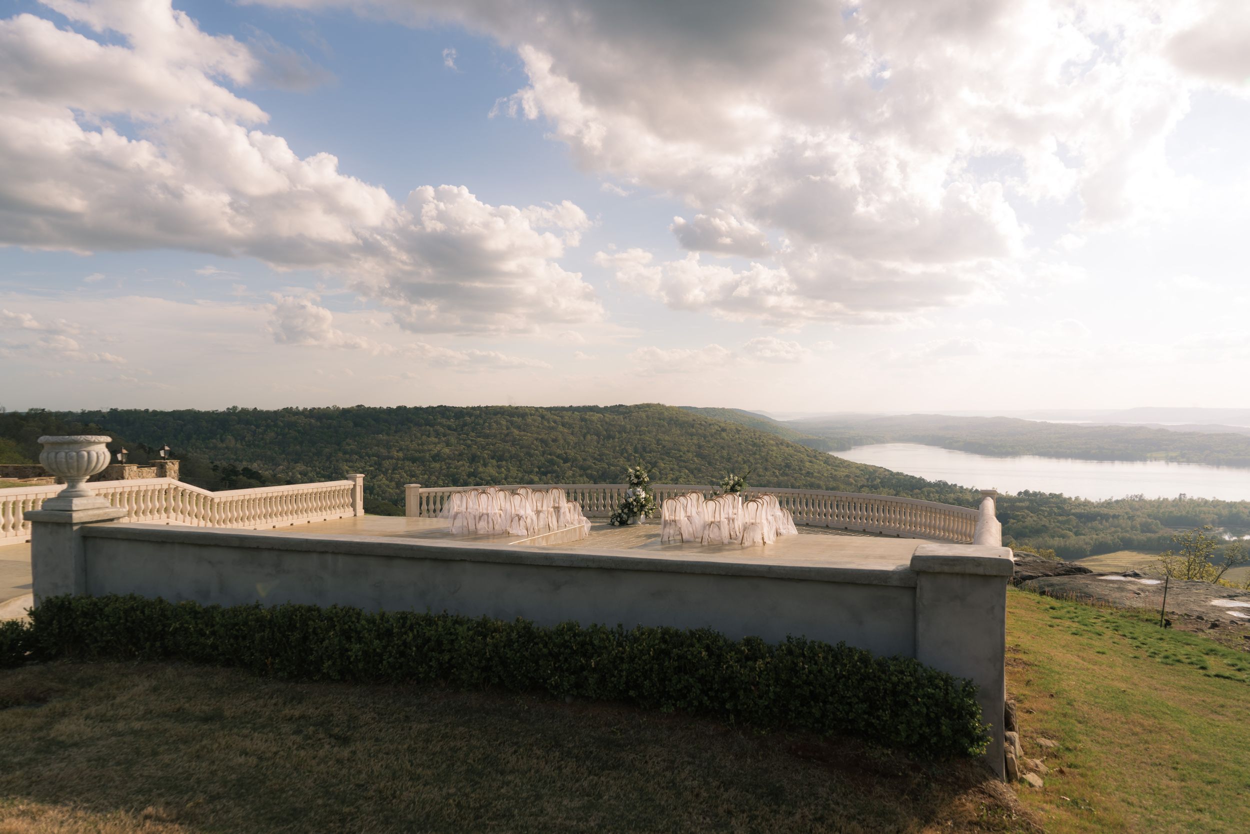Elegant terrace with fountains overlooking lush green hills and a river under a partly cloudy sky.