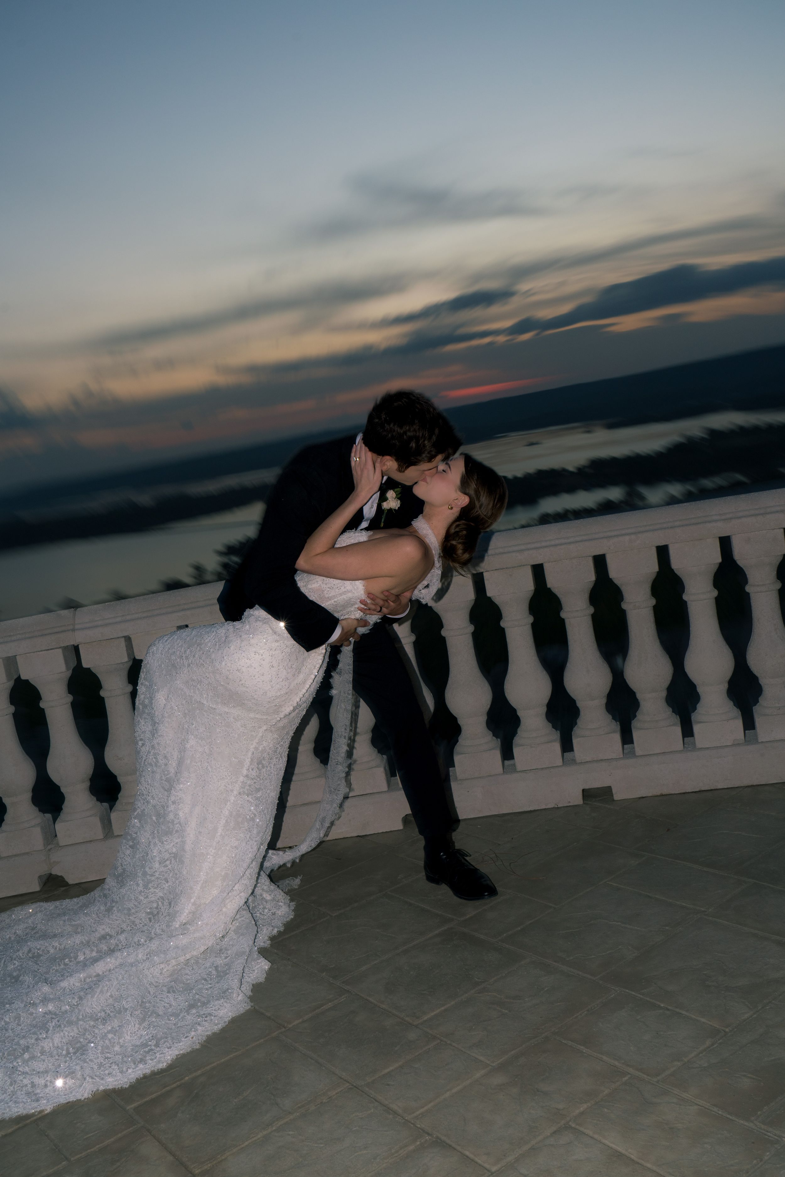 Bride and groom share a romantic dip kiss on a balcony at sunset with scenic waterfront views.