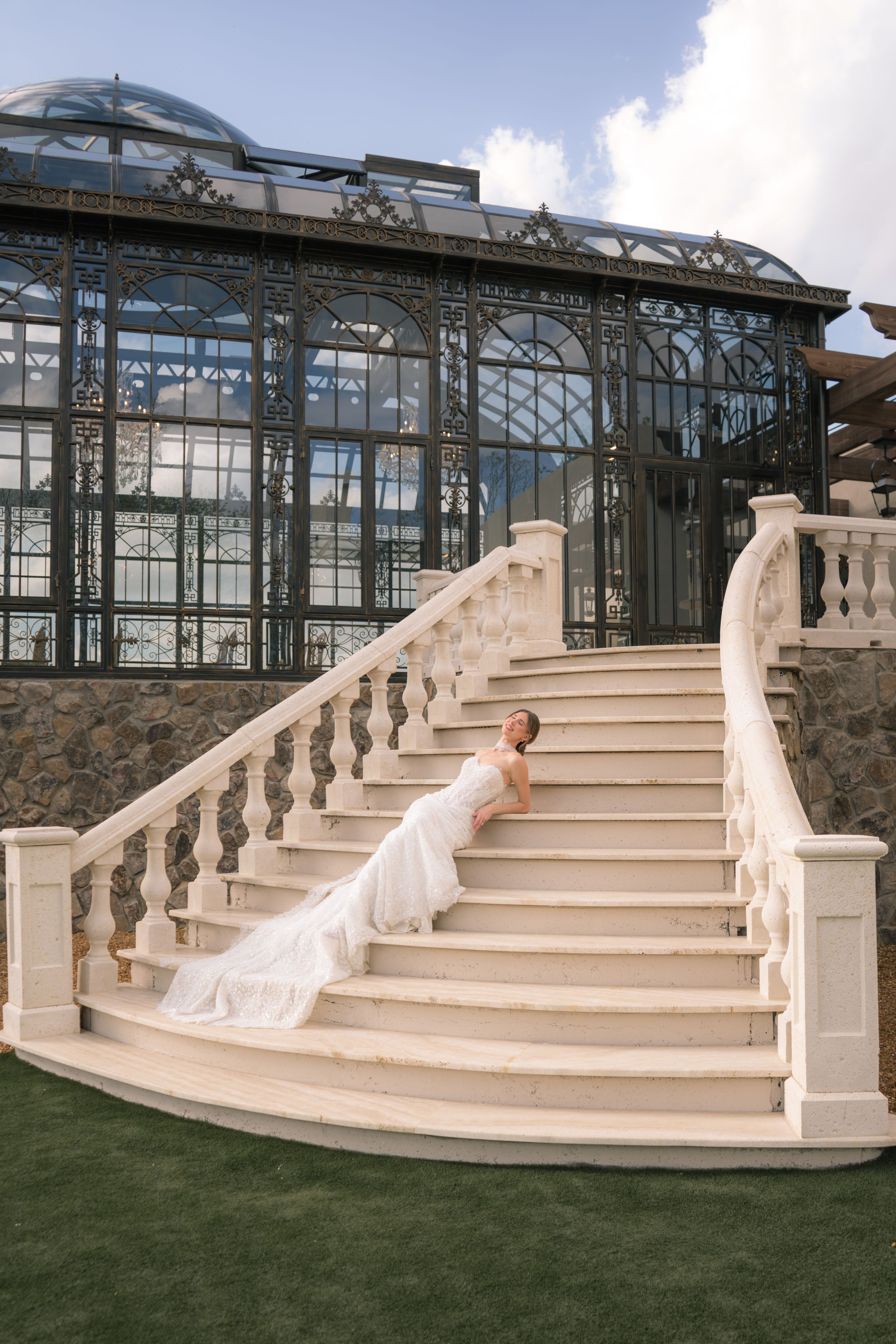 Bride in white gown posing on elegant white marble staircase outside a grand glass conservatory building.