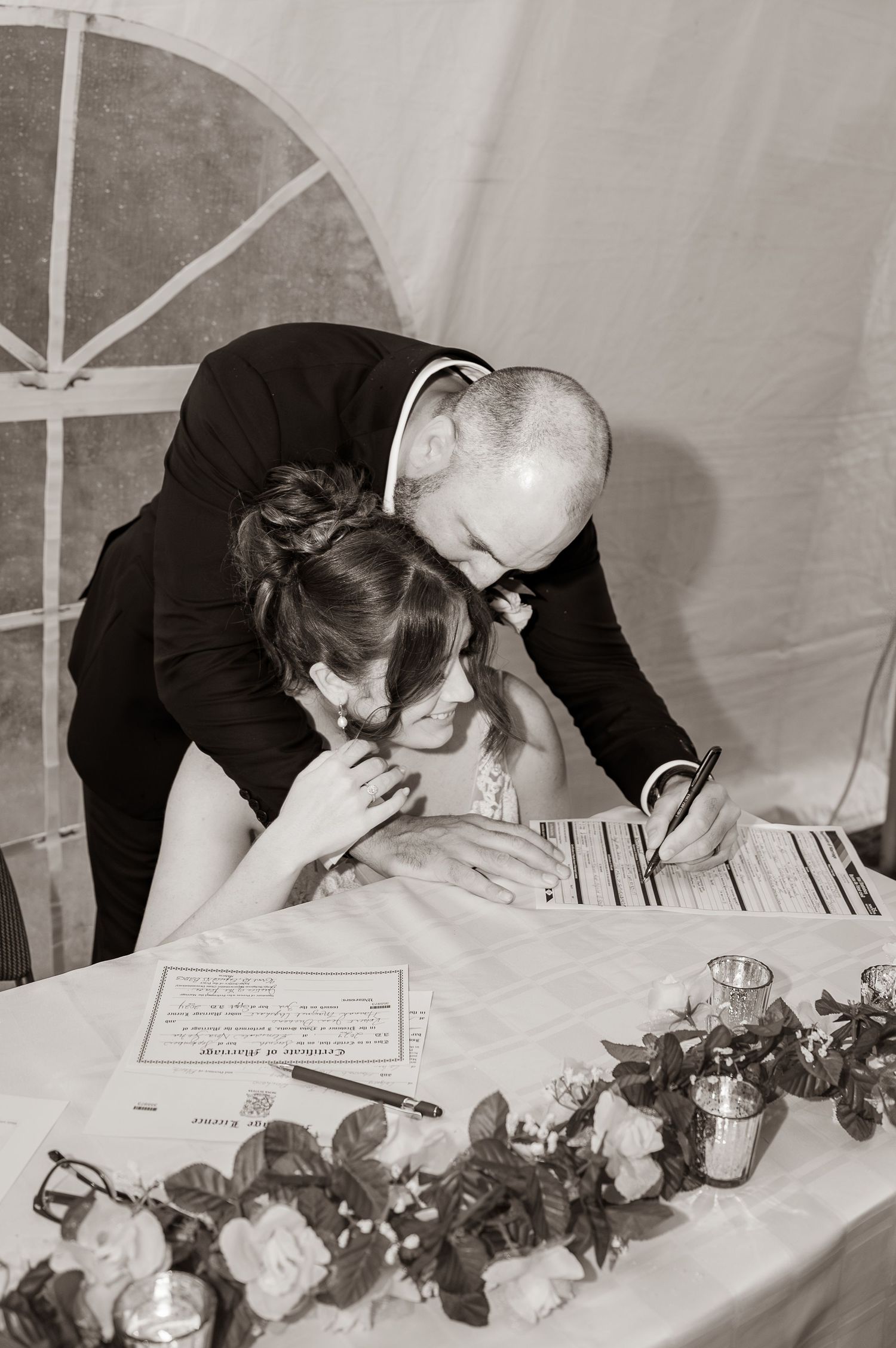 Newly married couple joyfully signing their marriage certificate during their wedding celebrations, while he kisses her on the forehead