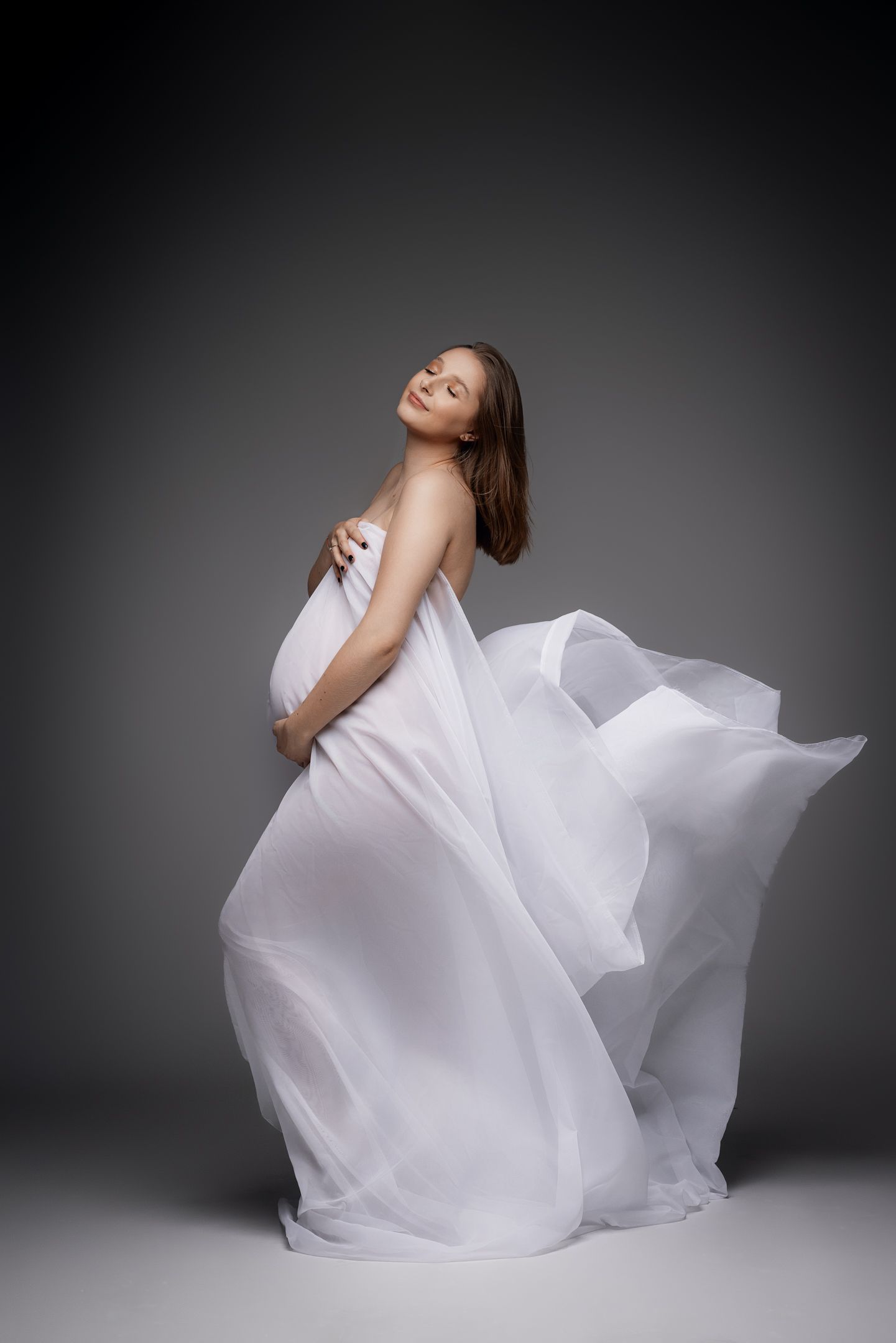 Pregnant woman in flowing white dress poses dramatically against dark background.