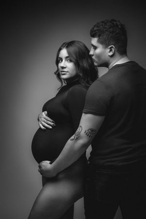 Artistic black and white maternity portrait of couple standing against dark backdrop.