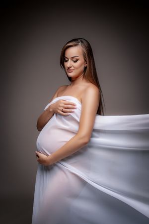 Elegant maternity portrait with flowing white fabric against dark studio backdrop.