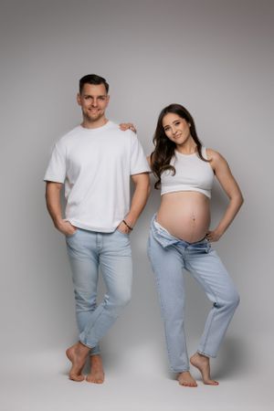 Casual maternity portrait of an expecting couple in matching white and denim against grey background.