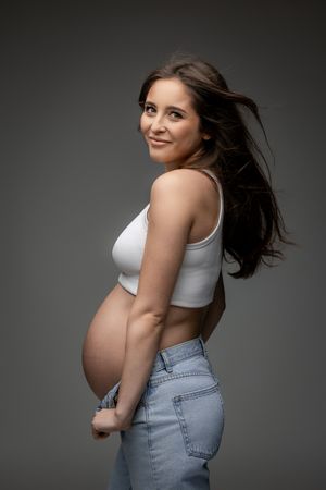 Side view maternity portrait of an expectant mother in casual denim against a gray backdrop.
