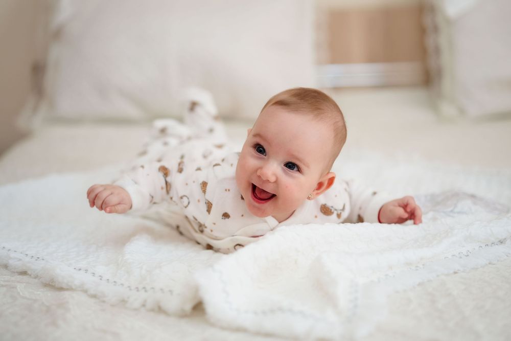 A baby in white clothing lies on soft white bedding with a joyful expression.