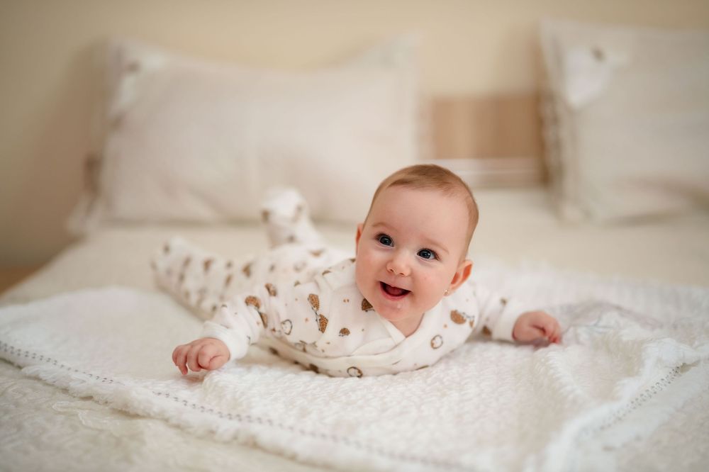 A baby wearing floral clothing lies on white bedding while smiling and playing with a soft blanket.