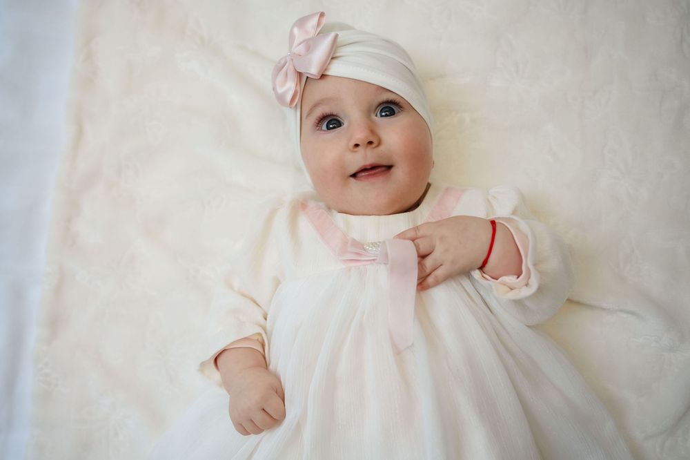 Baby wearing pink dress and white hat lies on white blanket smiling and playing in four sequential photos.