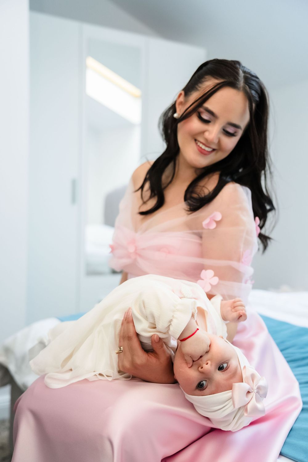 A mother in pink dress holds and plays with baby wearing white outfit on pink bedding in bright room.