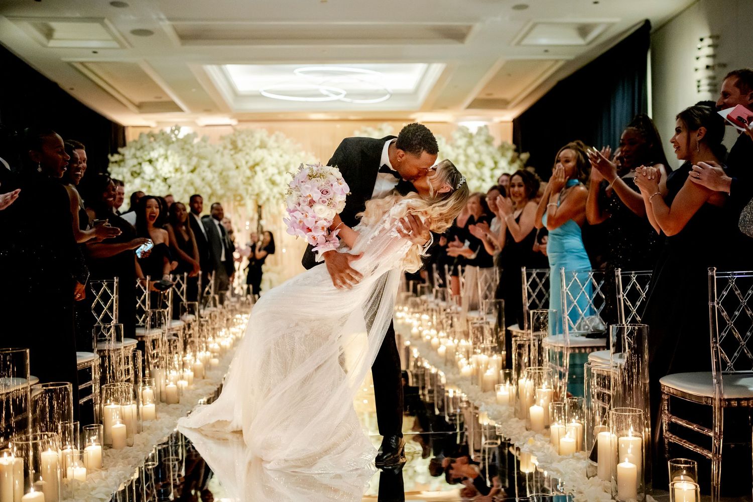 Groom dips and kisses his bride in a white gown down a candlelit wedding aisle as joyful guests clap and cheer on both sides.